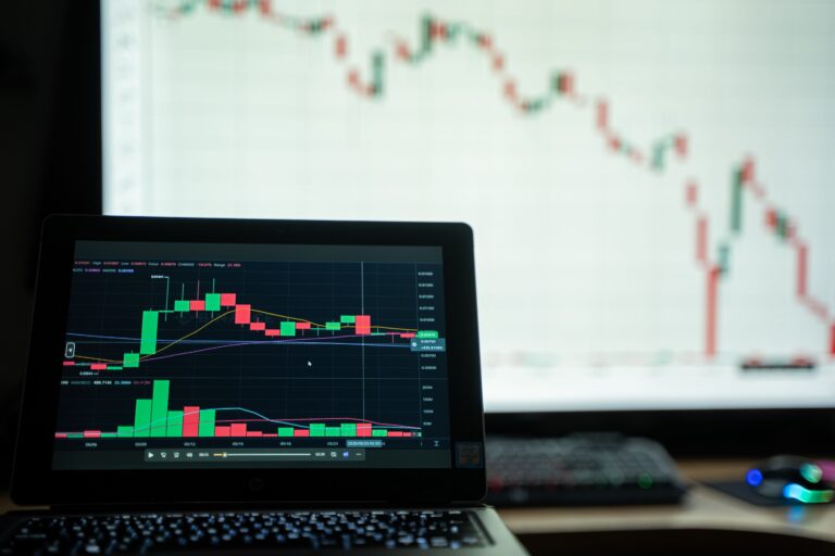 Laptop showing a candlestick chart in front of a larger screen with a longer-term price trend